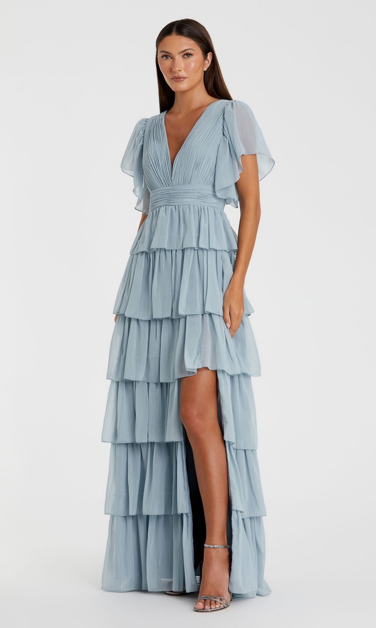 A woman wears the Mac Duggal 49595 Flutter-Sleeve Long Formal Dress—a light blue, tiered, floor-length gown with flutter sleeves and a deep V-neckline—standing against a plain white background.